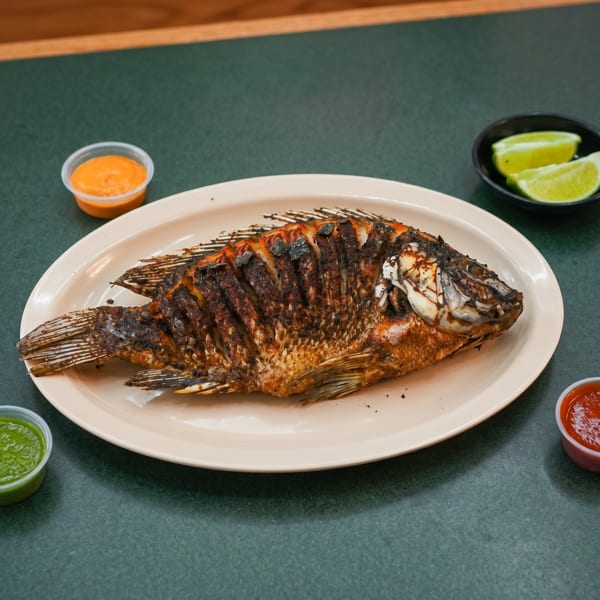 Whole fried fish on a white plate, with lime wedges and three dipping sauces on a green table.