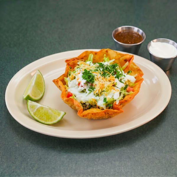 Taco salad in a crispy tortilla bowl with lime wedges, salsa, and sour cream.