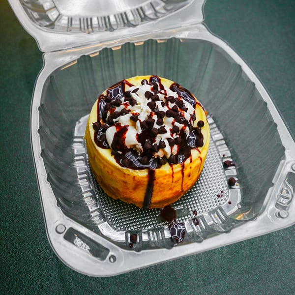Cheesecake with whipped cream, chocolate syrup, and chocolate chips in a clear to-go container.
