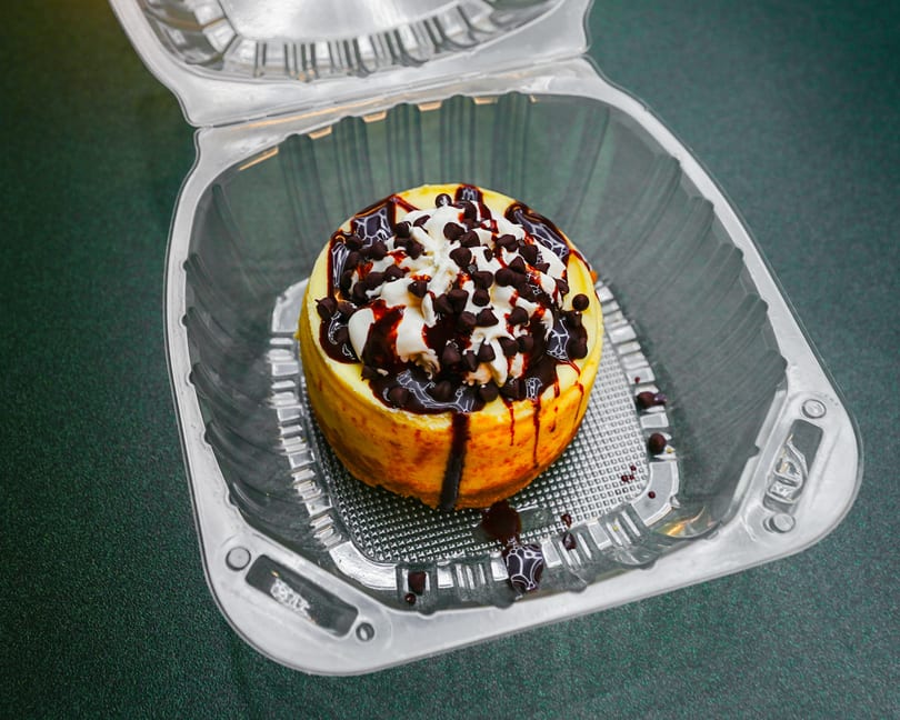 Cheesecake with whipped cream, chocolate syrup, and chocolate chips in a clear to-go container.