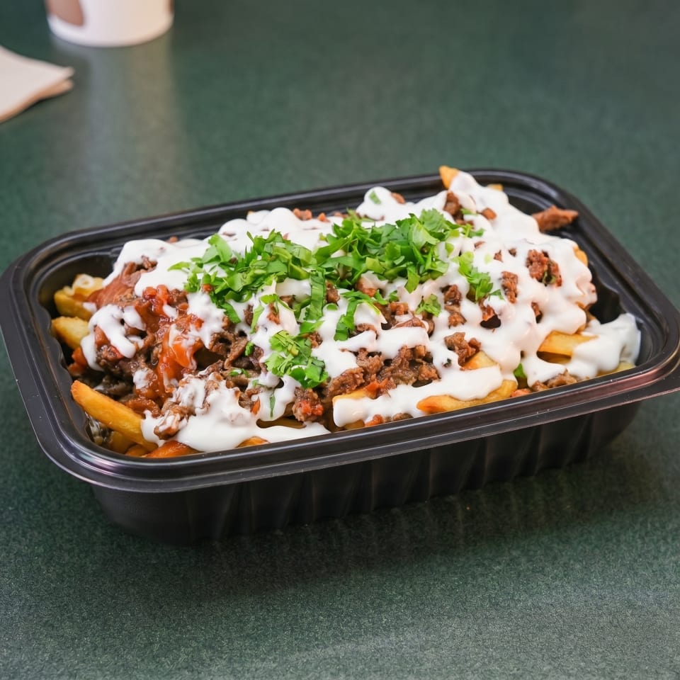 Loaded fries with ground meat, white sauce, and fresh green herbs in a black takeout container.