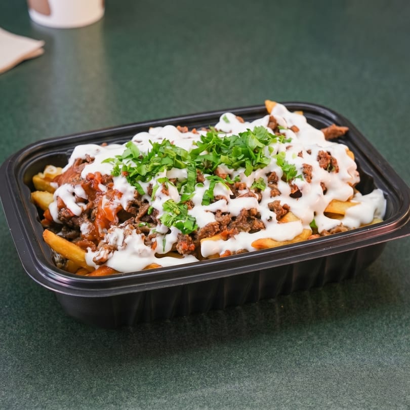 Loaded fries with ground meat, white sauce, and fresh green herbs in a black takeout container.