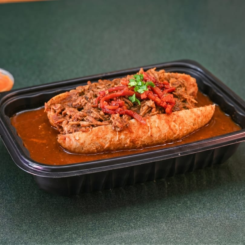 A black takeout container holds a large sandwich submerged in a rich brown sauce, topped with shredded meat, red peppers, and cilantro.
