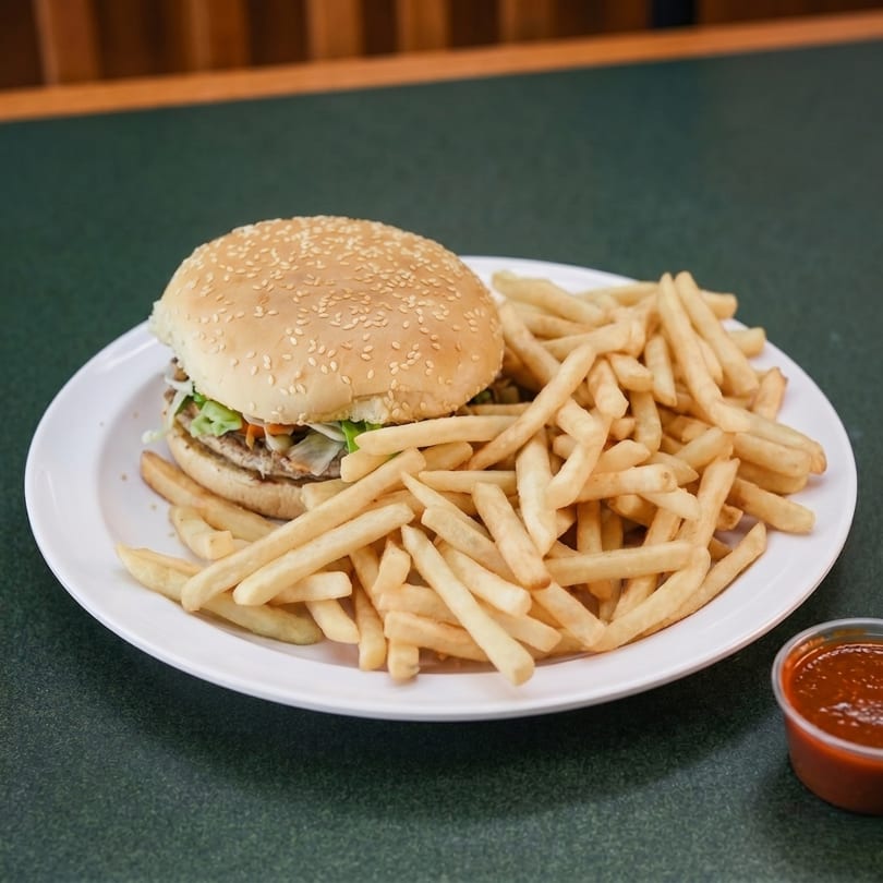 A sesame seed bun burger with lettuce and fries on a white plate with a side of sauce.