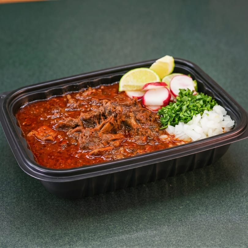 Shredded beef birria in a black takeout container with limes, radishes, cilantro, and onions.