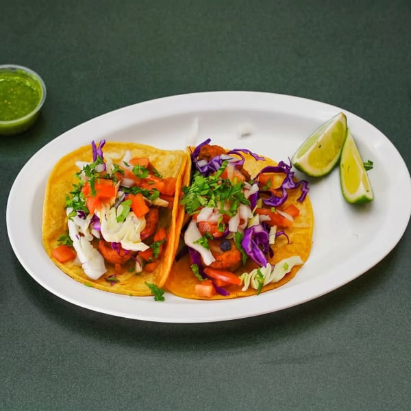 Two vibrant tacos with red cabbage, white cabbage, tomatoes, and cilantro, served with lime wedges and green sauce.