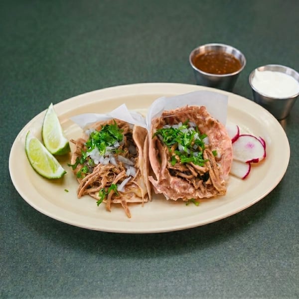 Two carnitas tacos with cilantro, onions, lime, and radishes, served with salsa and sour cream.